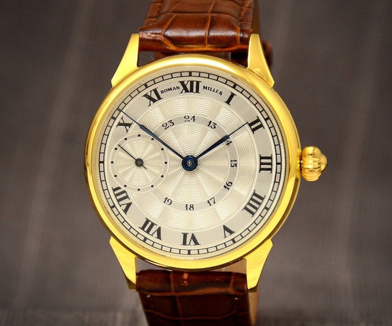 May include: A gold-toned wristwatch with a light-colored face, featuring Roman numerals and the text "ROMAN MILLER." The watch has a brown leather band and a smaller subdial. The watch hands are dark blue.