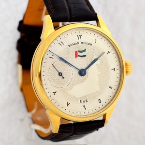 May include: A gold-toned wristwatch with a cream-colored face, featuring Arabic numerals and blue hands. The watch face displays the text "ROMAN MILLER" and the UAE flag. The watch has a dark brown leather strap.