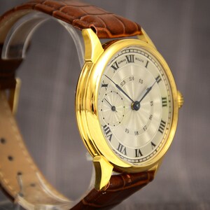 May include: A gold-toned wristwatch with a light-colored face and Roman numeral hour markers. The watch features a subdial and a date display. It has a brown leather band.