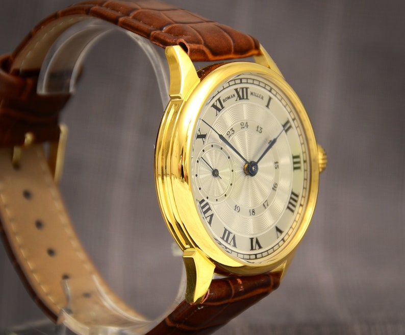 May include: A gold-toned wristwatch with a light-colored face and Roman numeral hour markers. The watch features a subdial and a date display. It has a brown leather band.