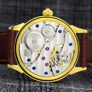 May include: A gold-toned wristwatch with a brown leather strap. The watch face reveals intricate mechanical components, including gears, screws, and jewels. The watch has a round face and a gold-colored crown.