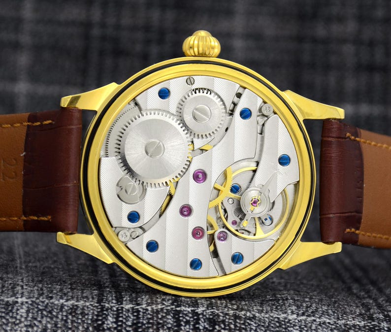 May include: A gold-toned wristwatch with a brown leather strap. The watch face reveals intricate mechanical components, including gears, screws, and jewels. The watch has a round face and a gold-colored crown.