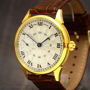 May include: A gold-toned wristwatch with a cream-colored face and Roman numeral hour markers. The watch features a brown leather band and a smaller subdial. The watch face reads "ROMAN MILLER".