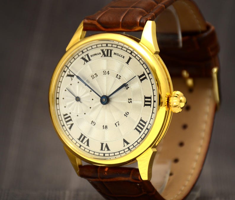 May include: A gold-toned wristwatch with a cream-colored face and Roman numeral hour markers. The watch features a brown leather band and a smaller subdial. The watch face reads "ROMAN MILLER".