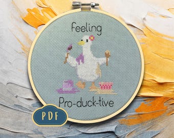 Puede incluir: Bordado de punto de cruz en un aro de madera con un pato de dibujos animados y las palabras "Feeling Pro-duck-tive". El pato es blanco con alas moradas y una flor en la cabeza, sosteniendo pinceles. Un icono PDF está en la esquina inferior izquierda.