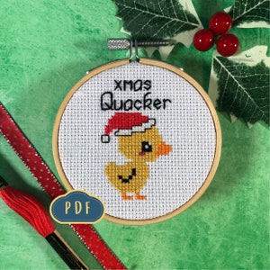 Puede incluir: Un patrón de punto de cruz con un pato amarillo que lleva un gorro de Papá Noel rojo. El patrón está enmarcado en un aro de madera e incluye el texto "xmas Quacker".