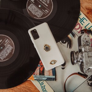 May include: A white Samsung Galaxy smartphone with a black camera lens and two gold-colored stickers on the back. The phone is lying on top of a vintage record and a book with the text "BILLY PRESTON SYREETA" and "is born".