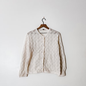 以下が含まれることがあります： 白いレースのトリムが付いたボタン留めの白いクロシェカーディガン。カーディガンは白い背景のウッドハンガーにかけられています。