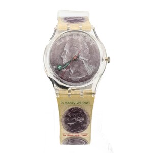 Puede incluir: Un reloj de pulsera de plástico transparente con una banda de color dorado. La esfera del reloj es una moneda de un cuarto de dólar estadounidense de color plateado con el texto "in money we trust" e "in time we trust" impreso en la banda.