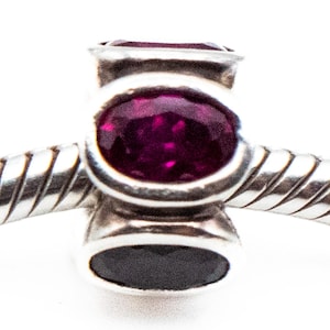 May include: A silver charm with a dark purple oval-shaped gemstone. The charm has a cylindrical shape with a smaller black oval at the bottom. The charm is attached to a silver, textured bracelet.