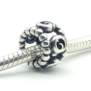 May include: A silver charm bead with a spiral design. The bead is round with a textured surface and is strung on a silver bracelet. The charm has a detailed, ornate pattern and is set against a white background.