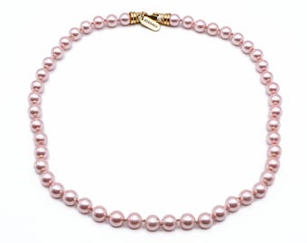 May include: A delicate pink pearl necklace with a gold clasp. The necklace features evenly spaced, round pearls. The clasp is gold-toned and has the word "KISSAKA" engraved on it. The necklace is displayed on a white background.