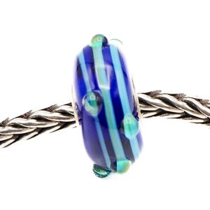 Puede incluir: Una cuenta de vidrio azul con rayas verticales en tonos azules y turquesas. La cuenta tiene pequeños acentos redondos de color turquesa y está ensartada en una cadena de plata. La cuenta es un elemento decorativo, probablemente para joyería o manualidades.