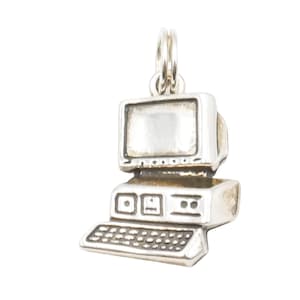 May include: A silver-colored charm in the shape of a vintage computer. The charm features a monitor, keyboard, and base, all in a detailed miniature design. The charm has a loop at the top for attaching to a bracelet or necklace. A retro tech-themed accessory.