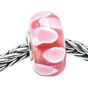 Puede incluir: Una cuenta de vidrio rosa y blanca con un patrón floral. La cuenta es cilíndrica y está ensartada en una cadena de plata. El color rosa es translúcido y el patrón blanco es opaco.