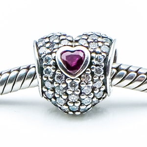 Può includere: Un ciondolo a forma di cuore in argento con un piccolo cuore rosso al centro, circondato da pietre di zircone cubico trasparenti.