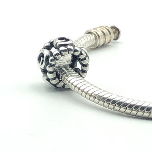 May include: A silver charm bead with a detailed, textured design, threaded onto a silver bracelet. The charm is round with a pattern of small, raised elements and open spaces. The bracelet has a snake chain design.