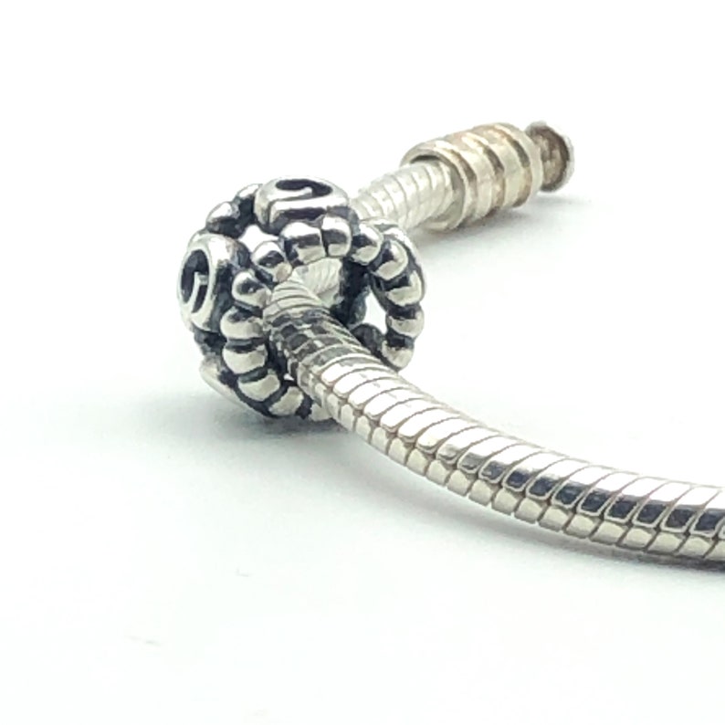 May include: A silver charm bead with a detailed, textured design, threaded onto a silver bracelet. The charm is round with a pattern of small, raised elements and open spaces. The bracelet has a snake chain design.