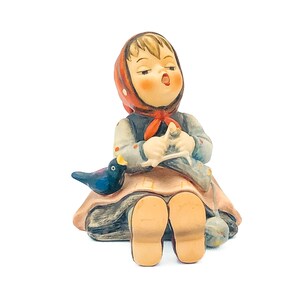 May include: A ceramic figurine of a seated girl wearing a red headscarf and a blue and gray outfit. She is knitting with a blue bird perched beside her. The figurine has a vintage aesthetic.