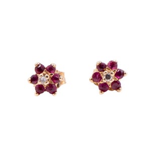 May include: A pair of gold stud earrings, each designed as a flower shape with six deep red ruby petals surrounding a single clear white diamond center stone. The metal setting is a warm yellow gold, providing a contrast to the gemstones.