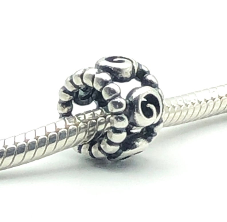May include: A silver charm bead with a spiral design. The bead is round with a textured surface and is strung on a silver bracelet. The charm has a detailed, ornate pattern and is set against a white background.