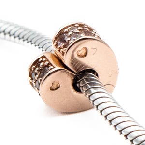 Puede incluir: Un charm de oro rosa con diseño de corazón, con pequeños cristales incrustados y un pequeño detalle de corazón. El charm está ensartado en una cadena de serpiente plateada, creando una joya.