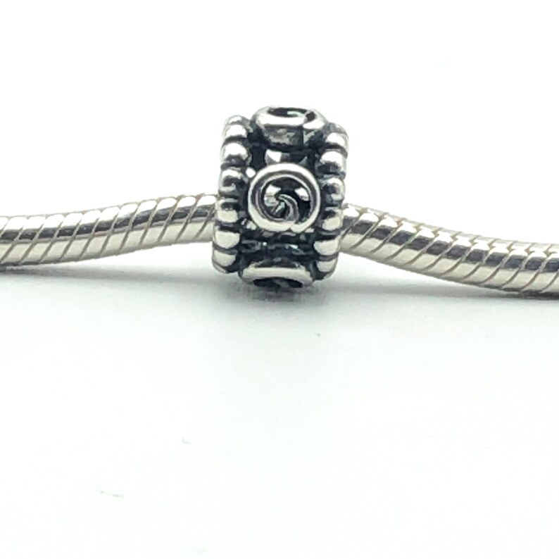 May include: A silver-toned charm bead with a spiral design in the center. The bead is cylindrical with a textured, beaded pattern around the edges. It is strung on a silver chain, set against a white background.