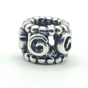 May include: A silver-colored charm with a cylindrical shape. The charm features a repeating pattern of rounded beads along the top and bottom edges. The sides display a stylized, swirling design. The charm is designed to be used with a bracelet or necklace.