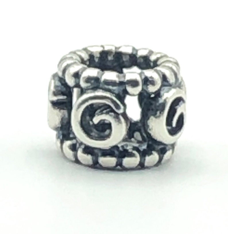 May include: A silver-colored charm with a cylindrical shape. The charm features a repeating pattern of rounded beads along the top and bottom edges. The sides display a stylized, swirling design. The charm is designed to be used with a bracelet or necklace.
