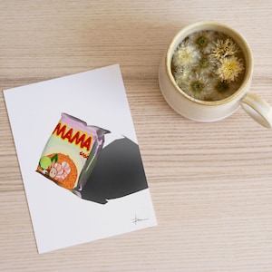 May include: A white card with a pink and orange packet of Mama instant noodles on it. The packet has a picture of prawns and vegetables. The card is on a light wood surface with a white mug of tea with white flowers in it.