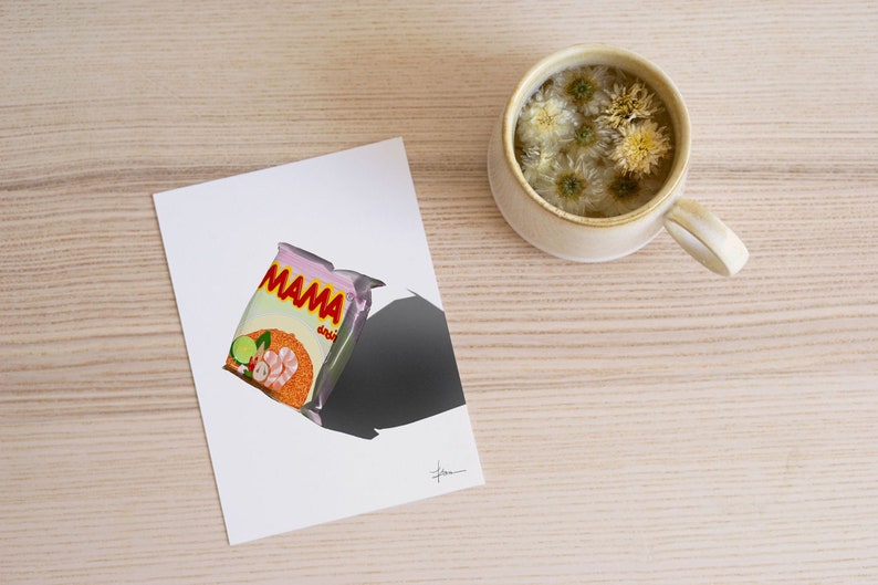 May include: A white card with a pink and orange packet of Mama instant noodles on it. The packet has a picture of prawns and vegetables. The card is on a light wood surface with a white mug of tea with white flowers in it.