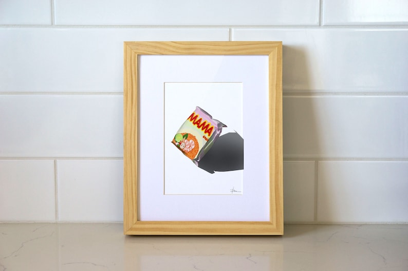 May include: A framed illustration of a pink and orange instant noodle packet with the text "MAMA" on it. The packet is floating in the air with a shadow below it.