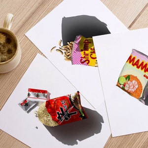 May include: Three different instant noodle packets on white paper. The packets are all different brands and flavours. One packet is red with white text, another is yellow with pink and white text, and the last is pink with white text. The packets are all slightly open and the noodles are spilling out.