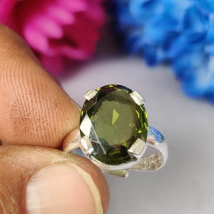 May include: A silver ring featuring a large, oval-cut, olive-green gemstone. The gemstone is set with four prongs. The ring is held up against a blurred background of pink and blue flowers.