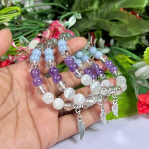 May include: Three beaded bracelets with clear, white, light blue, and purple stones. Each bracelet has a silver wing charm. The bracelets are displayed against a backdrop of green foliage and flowers.