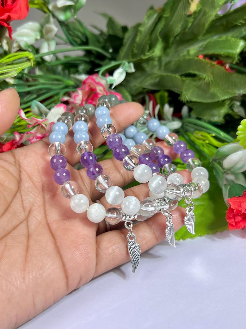 May include: Three beaded bracelets with clear, white, light blue, and purple stones. Each bracelet has a silver wing charm. The bracelets are displayed against a backdrop of green foliage and flowers.