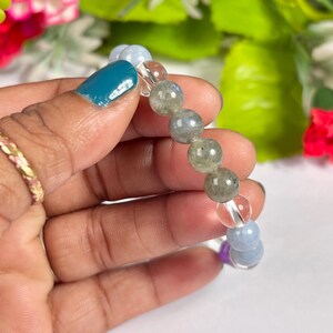 May include: A beaded bracelet featuring a mix of colorful gemstones. The bracelet includes clear, blue, green, and purple beads. The bracelet is held against a backdrop of green and pink flowers.