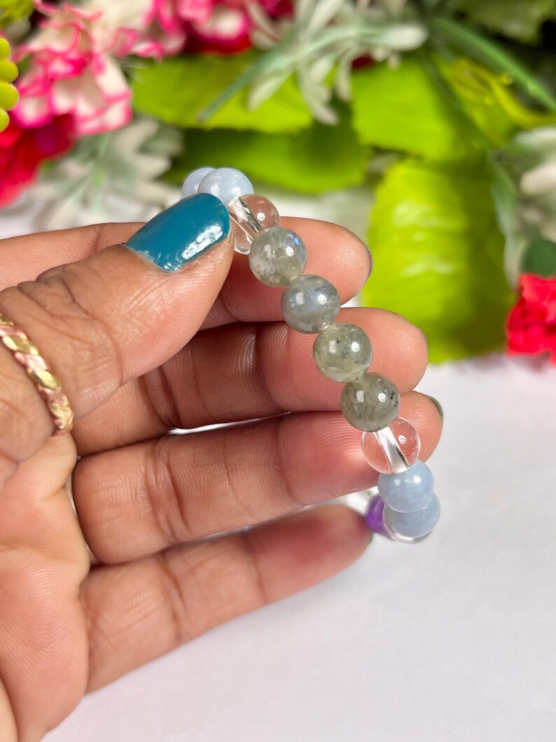 May include: A beaded bracelet featuring a mix of colorful gemstones. The bracelet includes clear, blue, green, and purple beads. The bracelet is held against a backdrop of green and pink flowers.