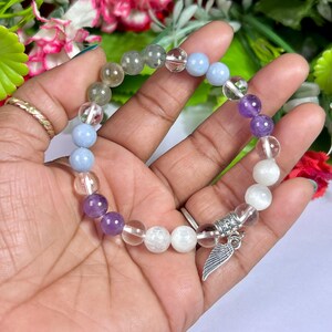 May include: A beaded bracelet featuring a variety of colorful gemstones, including clear, purple, blue, and white beads. A silver charm with a wing design hangs from the bracelet. The bracelet is held against a backdrop of flowers.