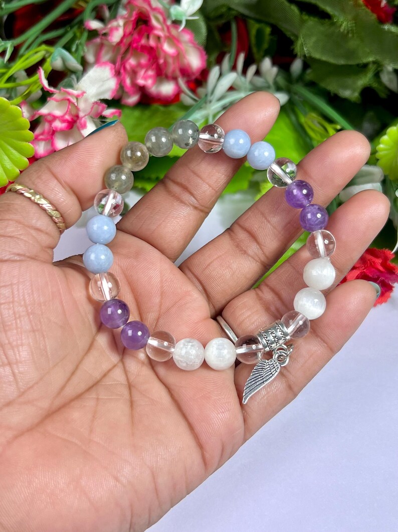 May include: A beaded bracelet featuring a variety of colorful gemstones, including clear, purple, blue, and white beads. A silver charm with a wing design hangs from the bracelet. The bracelet is held against a backdrop of flowers.