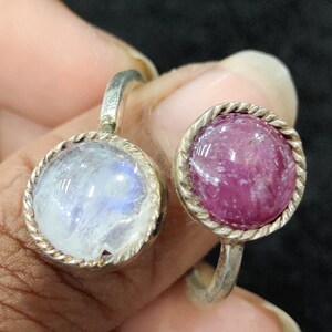 May include: Two silver rings with gemstone settings. The ring on the left has a white moonstone, and the ring on the right has a pink ruby.