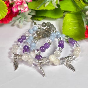 May include: Four beaded bracelets with clear, purple, light blue, and gray beads. Each bracelet has a silver charm with a wing design. The bracelets are displayed on a white surface with a floral background.