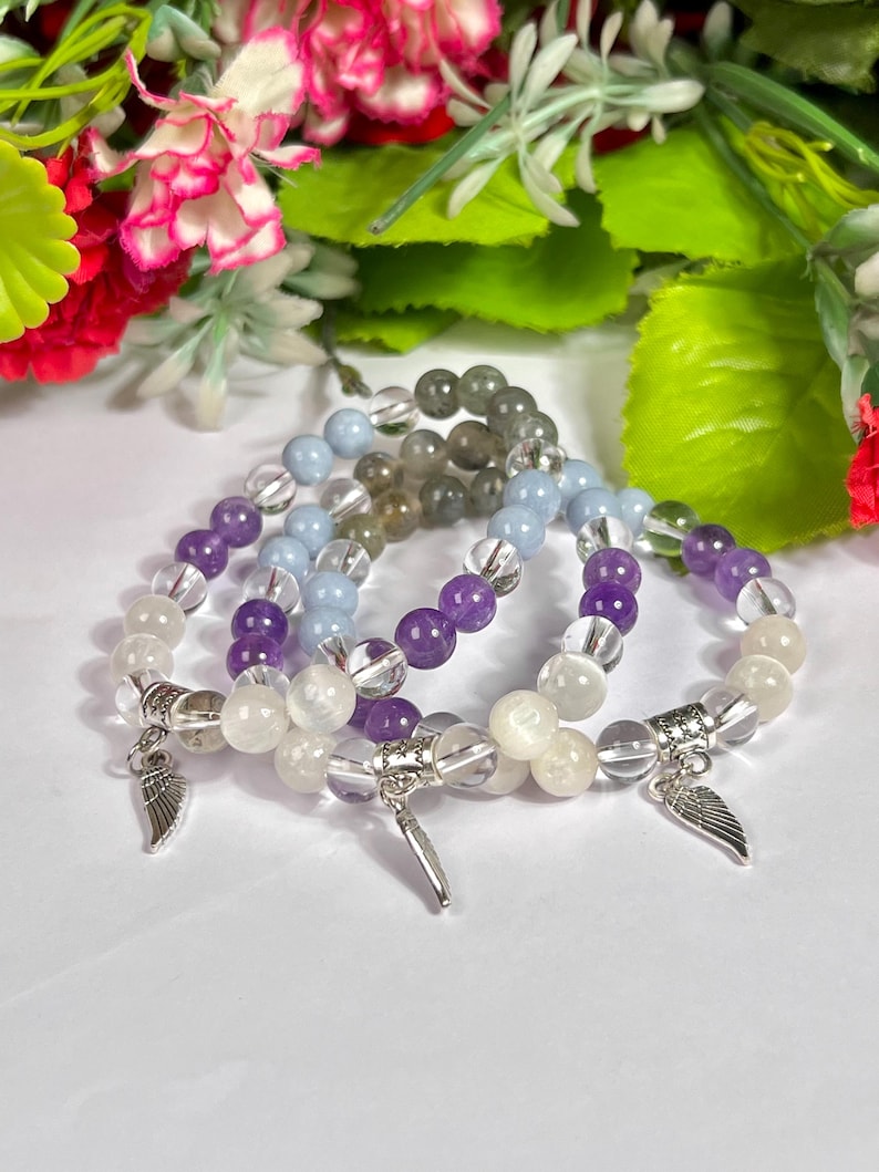 May include: Four beaded bracelets with clear, purple, light blue, and gray beads. Each bracelet has a silver charm with a wing design. The bracelets are displayed on a white surface with a floral background.