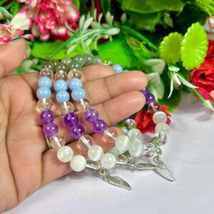 May include: Three beaded bracelets with clear, light blue, purple, and white beads. Each bracelet has a silver wing charm. The bracelets are held in a hand, with a blurred floral background.