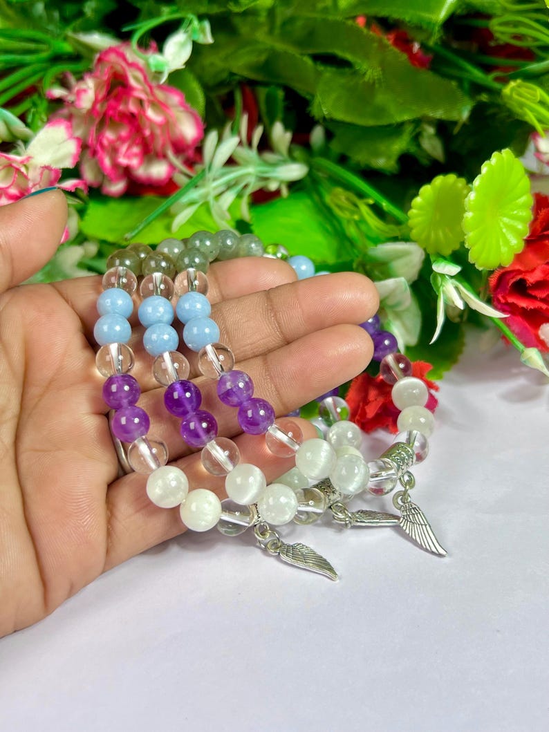 May include: Three beaded bracelets with clear, light blue, purple, and white beads. Each bracelet has a silver wing charm. The bracelets are held in a hand, with a blurred floral background.