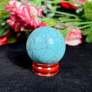 May include: A turquoise sphere with black veining sits on a small, round, red wooden stand. The sphere is a vibrant blue-green color. The background features pink and red flowers.
