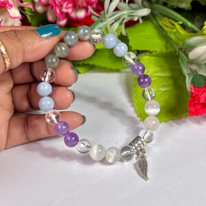 May include: A beaded bracelet featuring a mix of clear, purple, blue, and gray gemstone beads. A silver charm with a wing design hangs from the bracelet. The bracelet is held against a background of flowers.