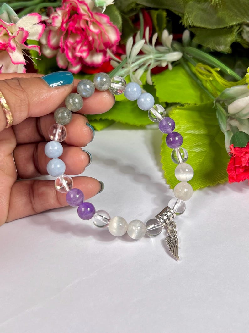 May include: A beaded bracelet featuring a mix of clear, purple, blue, and gray gemstone beads. A silver charm with a wing design hangs from the bracelet. The bracelet is held against a background of flowers.