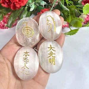 Könnte beinhalten: Vier ovale, durchscheinende weiße Selenitsteine, von denen jeder ein einzigartiges goldfarbenes Symbol oder ein japanisches Kanji-Schriftzeichen auf der Oberfläche eingraviert hat. Die Steine haben eine glatte, polierte Textur.