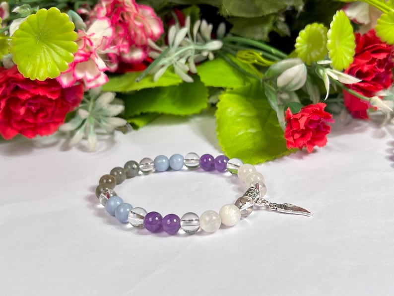 May include: A beaded bracelet featuring a variety of colorful gemstones, including clear, purple, blue, and gray beads. The bracelet has a silver charm and is set against a backdrop of flowers.